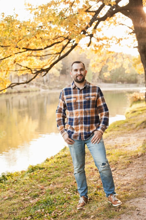 Preview: a guy in checkered shirt with beard smiling, standing alone in autumn park by the lake near tree