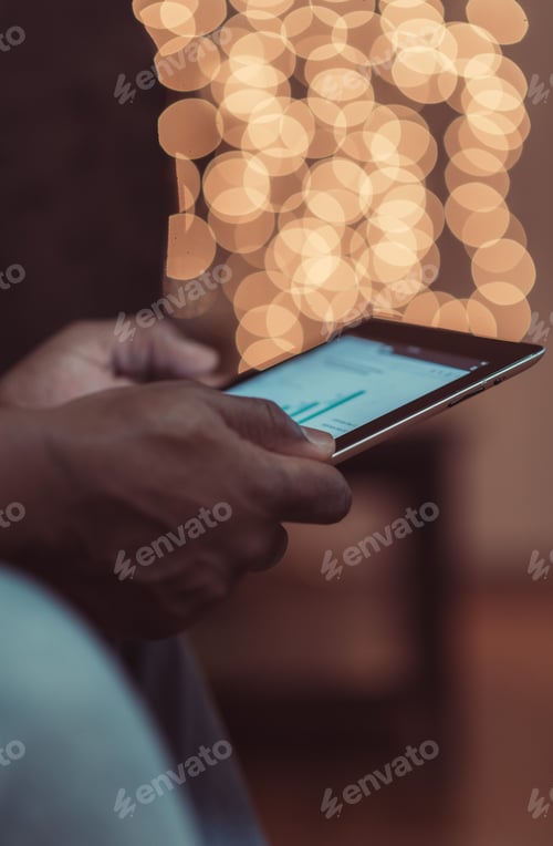 Preview: African American man holding a tablet with bar graphs displayed and festive Christmas lights in back