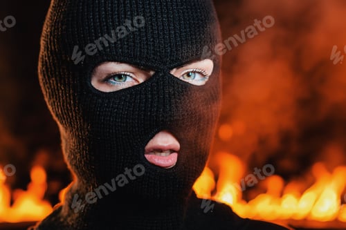 Preview: Portrait of young woman in black balaclava against backdrop of a blazing night fire.