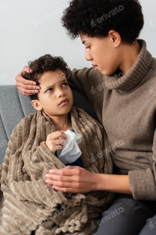 Preview: Portrait of little boy and beautiful mother holding thermometer sitting together on sofa at home