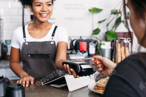 Preview: Smiling barista in apron holding POS terminal while customer paying by card