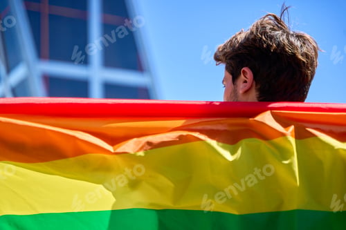 Preview: A man holding a rainbow flag in front of a building. The flag is large and colorful.