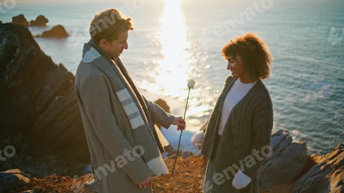 Preview: Lovely pair dating sunset sea cliffs closeup. Enamoured man bringing flower