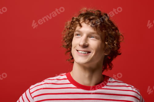 Preview: Stylish young man with curly hair smiles in a vibrant red studio setting