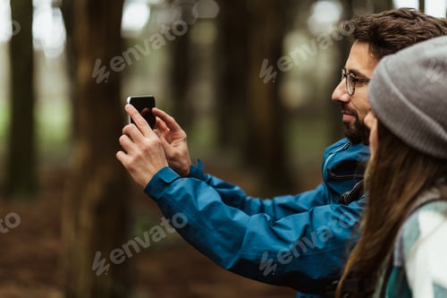 Preview: Cheerful young caucasian couple in jackets in forest enjoy travel vacation, make photo on smartphone