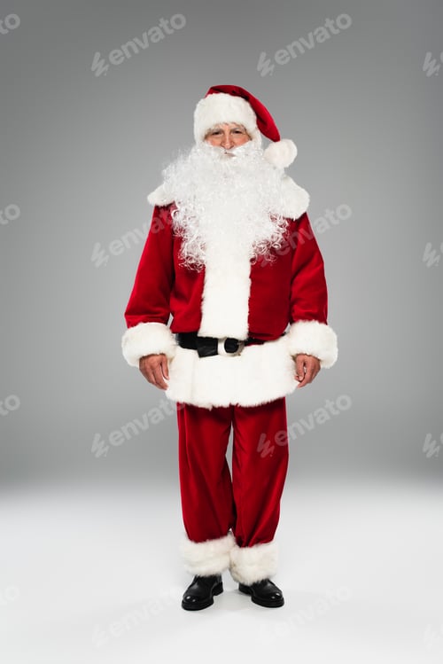 Preview: Full length of bearded father christmas looking at camera while standing on grey