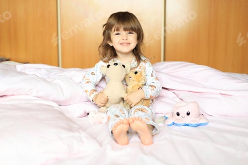 Preview: Happy cute little girl playing with children's toys in the bed at home. Light and airy