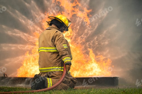 Preview: Two firefighters kneel in position while directing a powerful water stream at a large blaze