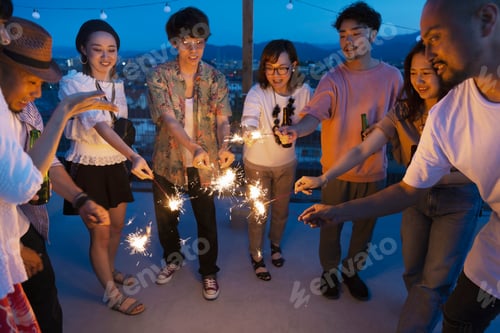 Preview: Group of young Japanese men and women with sparklers on a rooftop in an urban setting.
