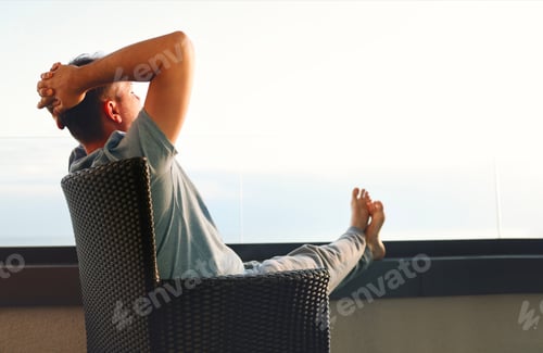 Preview: Young man in gray casual clothes sits on wicker chair and put his hands behind his head.