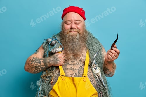 Preview: Satisfied handsome bearded seafarer stands with closed eyes holds smoking pipe dressed in yellow ove