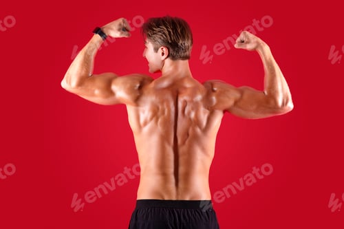 Preview: Young muscular sportsman standing with his naked back to camera and showing biceps on red studio