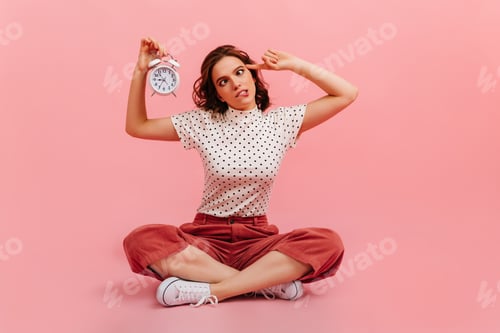 Preview: Carefree girl showing time on pink background. Funny young woman posing with clock.