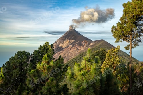 Preview: Scenic view of an active volcano emitting smoke, surrounded by lush green trees under a clear sky.