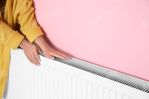 Preview: Woman warming hands on heating radiator near color wall