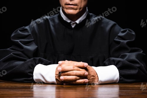 Preview: cropped view of judge in judicial robe sitting at table with clenched hands isolated on black