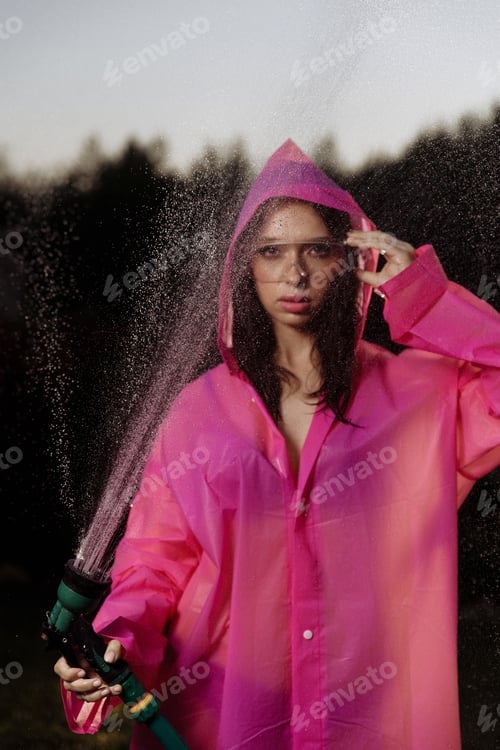 Preview: A glamour woman in a pink raincoat is spraying water from a hose