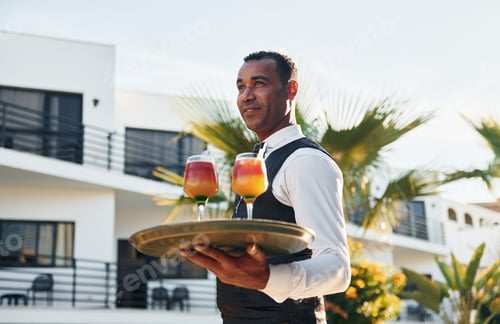 Preview: Holds cocktails. Black waiter in formal clothes is at his work outdoors at sunny daytime