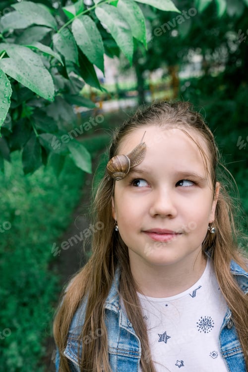 Preview: A young girl looks at a snail on her face in the green of the trees