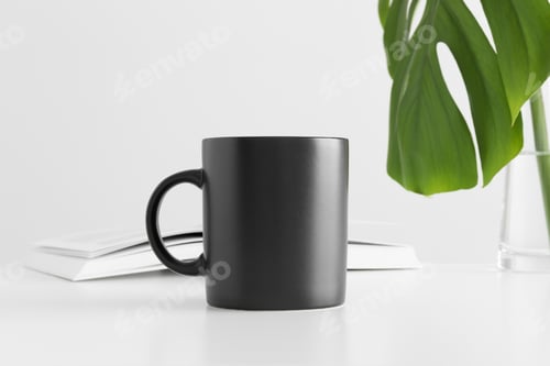 Preview: Black mug mockup with a monstera leaf in a vase and a book on a white table.
