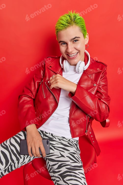 Preview: Positive carefree punk girl with dyed green hair poses on chair smiles gladfully uses smartphone and