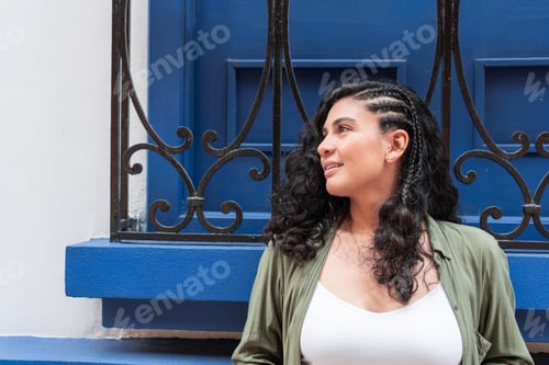 Preview: Stylish woman with braided hair standing by a blue wall with decorative ironwork.