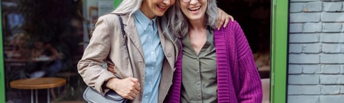 Preview: Pretty senior Asian lady hugs friend in purple jacket standing near cafe on street