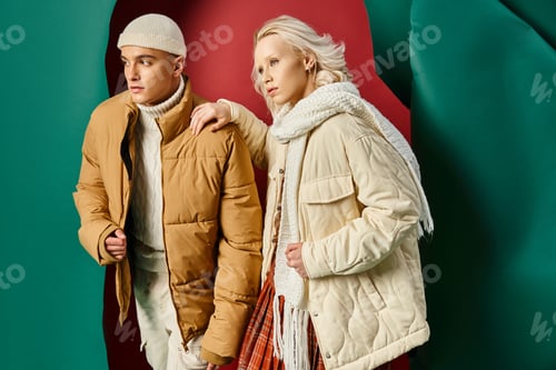 Preview: stylish man in beanie and puffer jacket posing with blonde woman on red with turquoise backdrop