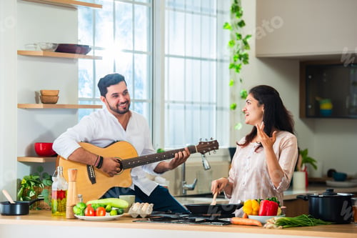 Preview: Asian Indian happy couple having fun while cooking food in kitchen, husband plays acoustic guitar