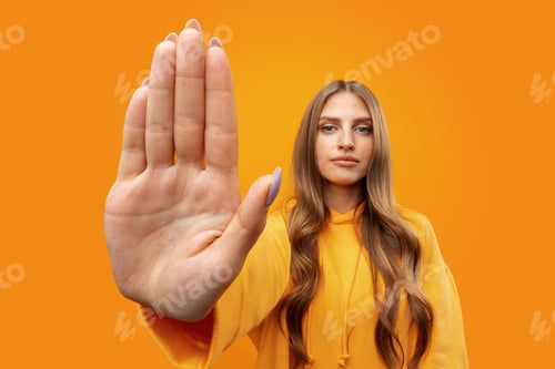 Preview: Young woman standing with outstretched arm showing stop sign against yellow background