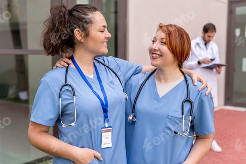 Preview: Two female nurses in blue scrubs, embraced and smiling, standing outdoors with doctor in background