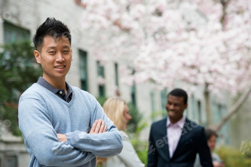Preview: A man with his arms folded and two people talking in the background under the trees in blossom.