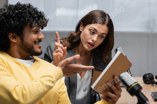 Preview: amazed podcaster showing idea sign while looking in notebook near cheerful indian colleague