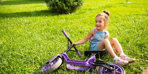 Preview: Cheerful girl sitting resting on the grass in a park near to the purple running bike in summer time.