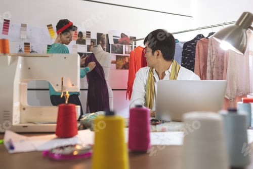 Preview: Asian male fashion designer talking with Mixed-race female designer while working on laptop