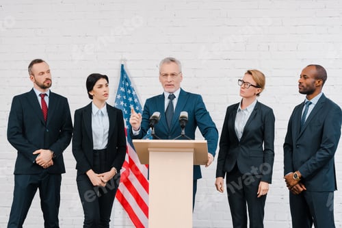 Preview: bearded speaker in glasses pointing with finger near microphones and multicultural diplomats