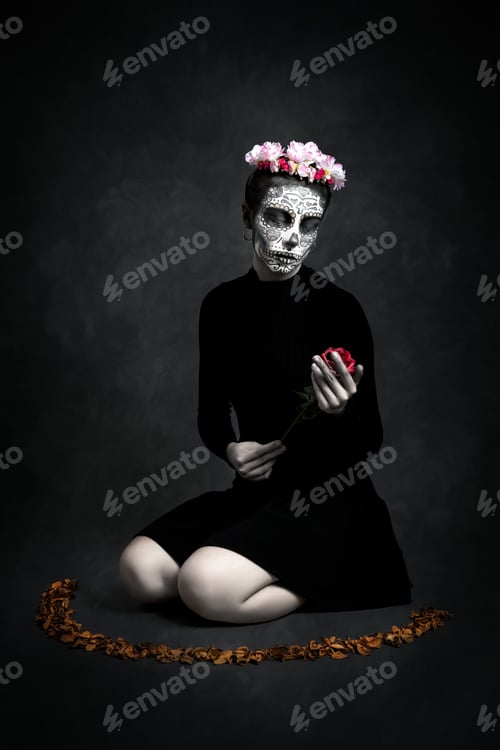 Preview: Painted Catrina woman and flower crown on her knees with a flower in her hand on dark background