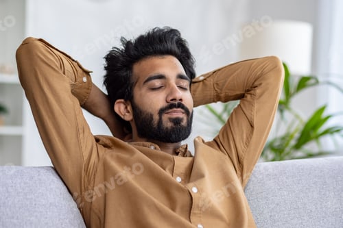 Preview: Relaxed young man with beard enjoying a moment of peace on cozy sofa with hands behind head, eyes