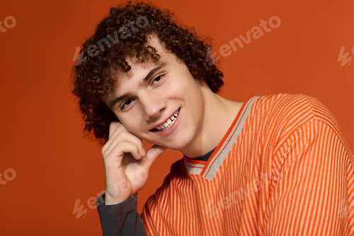 Preview: Curly-haired teenager in casual outfit smiles against bright orange backdrop