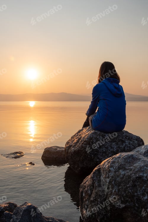 Preview: Girl looking sunset on sea, lake. Woman sitting on rocks. Lonely person. Girl alone outside