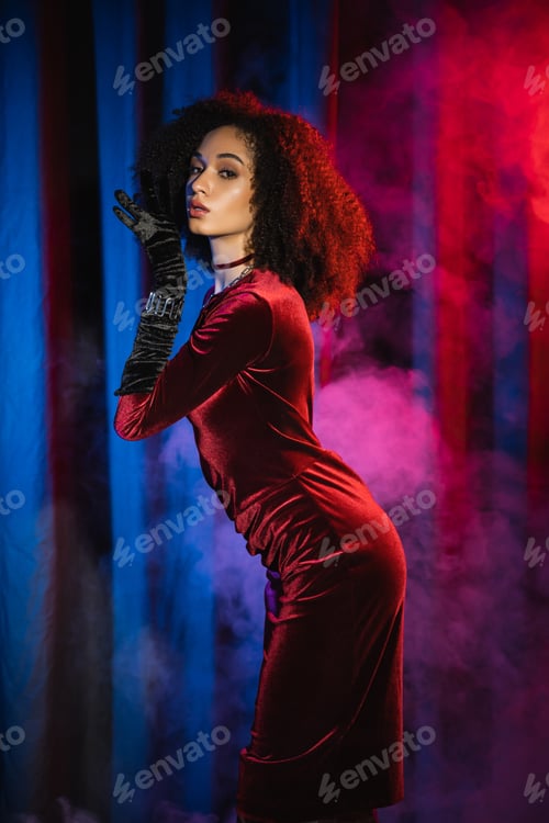 Preview: Stylish african american woman in velvet dress and glove posing near background with blue light