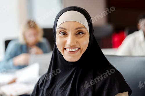 Preview: Young muslim woman smiling while working in busy office looking at camera