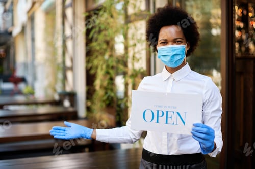 Preview: Happy black waitress holding open sign while wearing face mask and gloves at outdoor cafe.