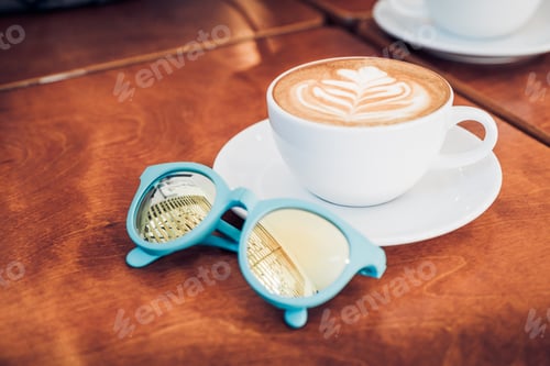 Preview: Close up white coffee cup with heart shape latte art and sunglasses on wood table at cafe..