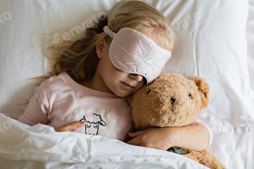 Preview: Cute little Caucasian girl with blonde hair wearing blindfold and sleeping in bed with teddy bear