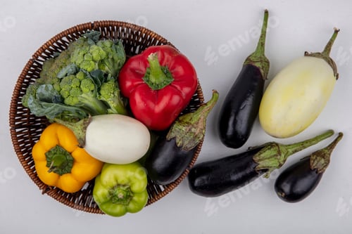 Preview: top view colored bell peppers with broccoli in basket with white and black eggplants on white