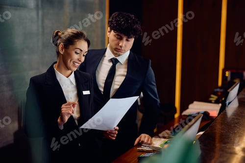 Preview: Happy concierge and her coworker analyzing paperwork at hotel reception desk.