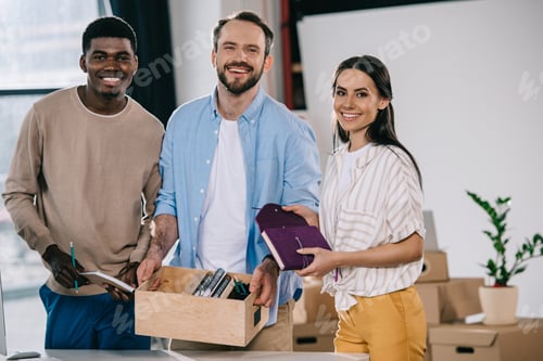 Preview: happy multiethnic colleagues with cardboard box and office supplies smiling at camera in new office