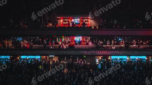 Preview: Balconies crowded with people and lit with neon lights and cast with dark shadows at a concert