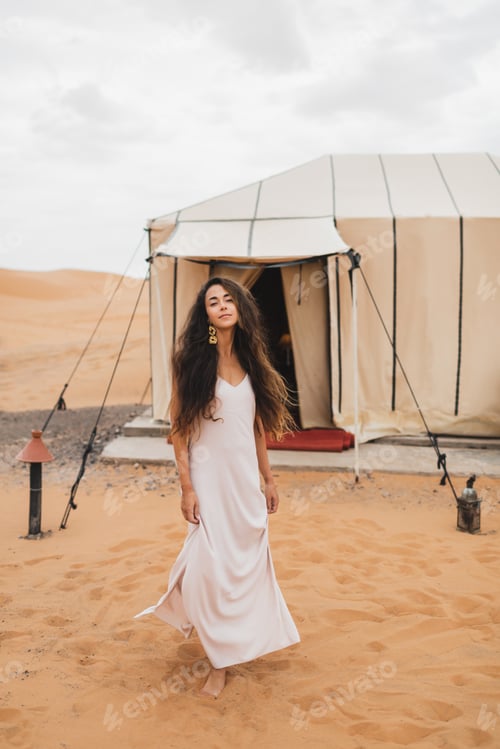 Preview: Portrait of beautiful oriental young brunette woman in white dress. Glamping tent in moroccan desert